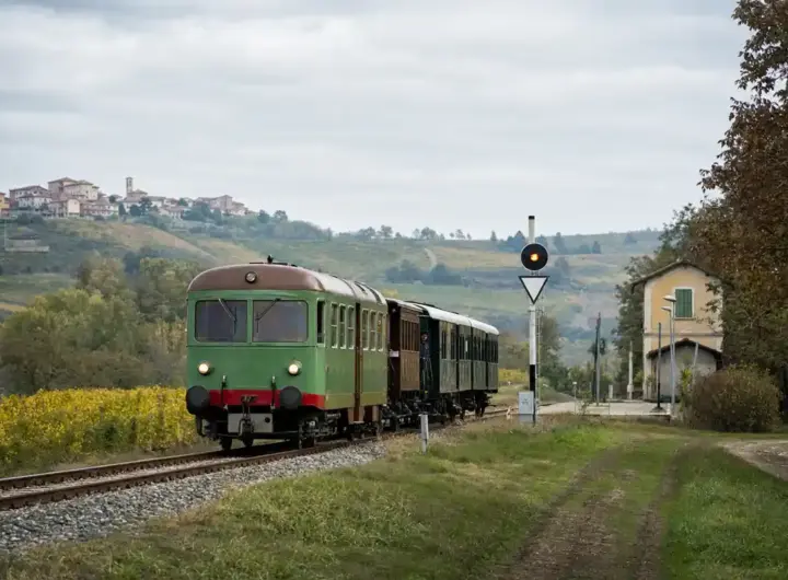 Treno storico verde su binari di campagna tra colline e vigneti, vicino a una piccola stazione