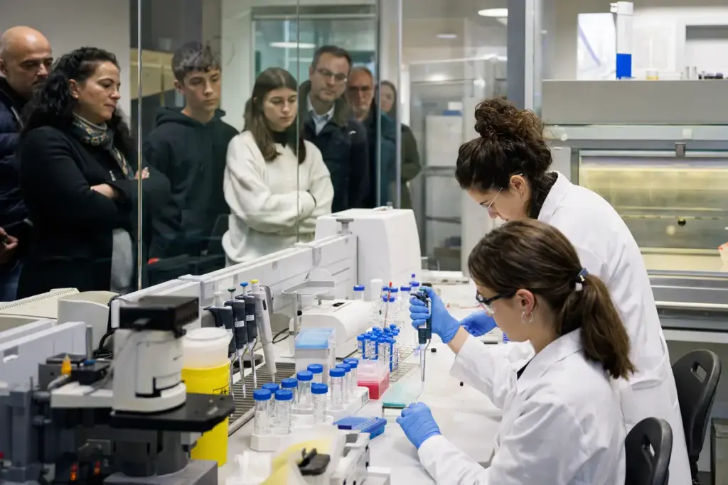 Ricercatrici in camice e guanti lavorano al banco con provette e pipette, mentre cittadini osservano dietro un vetro