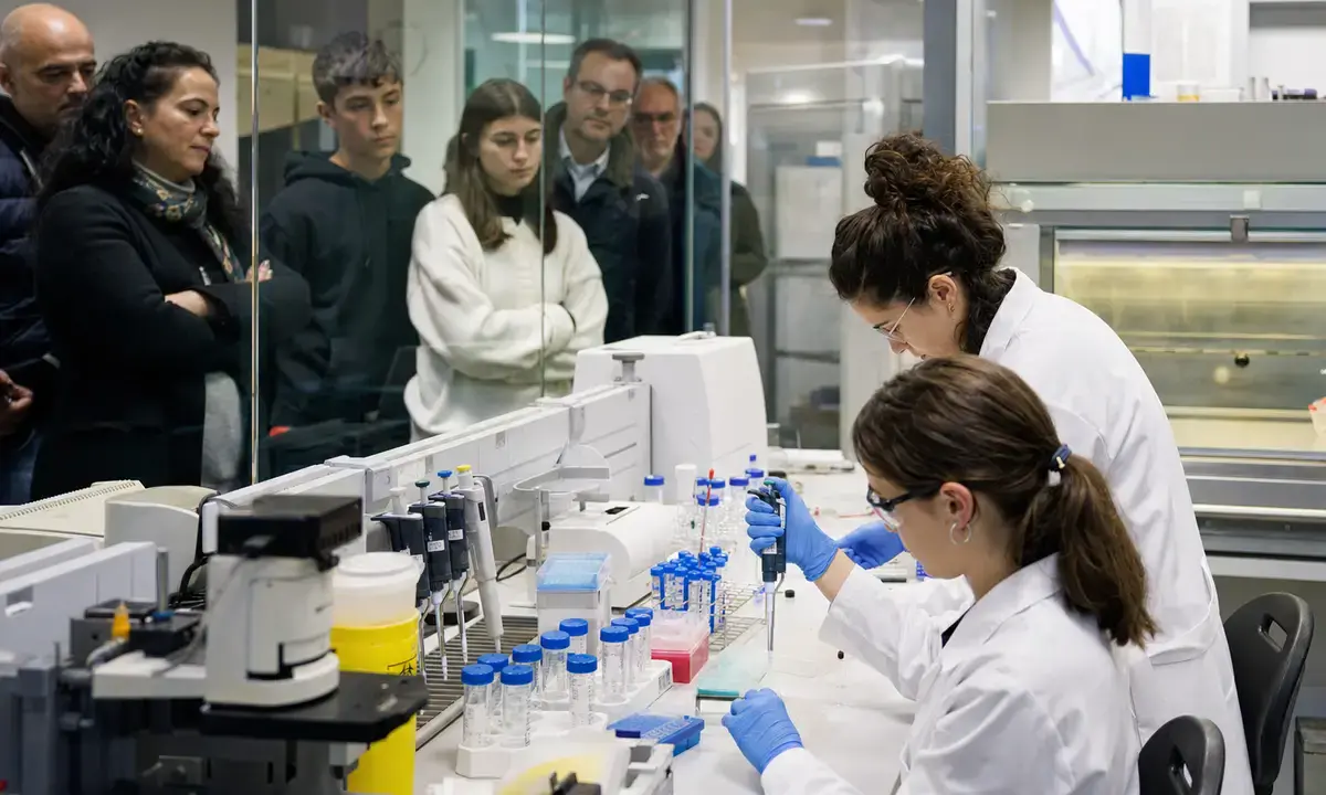 Ricercatrici in camice e guanti lavorano al banco con provette e pipette, mentre cittadini osservano dietro un vetro