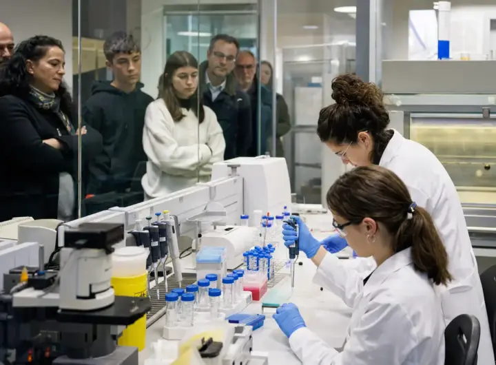 Ricercatrici in camice e guanti lavorano al banco con provette e pipette, mentre cittadini osservano dietro un vetro