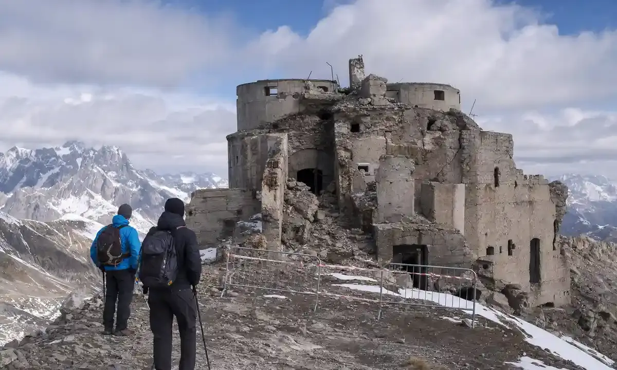 Due escursionisti davanti al Forte dello Chaberton in rovina, con transenne e nastro di divieto sulla vetta alpina