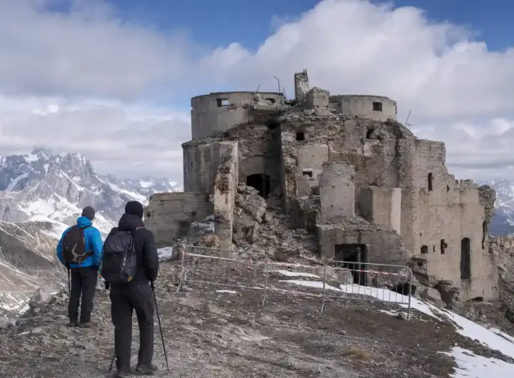 Due escursionisti davanti al Forte dello Chaberton in rovina, con transenne e nastro di divieto sulla vetta alpina