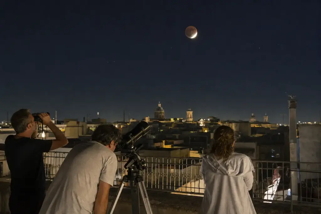 Persone su una terrazza osservano la Luna parzialmente eclissata con binocolo e telescopio sopra i tetti della città