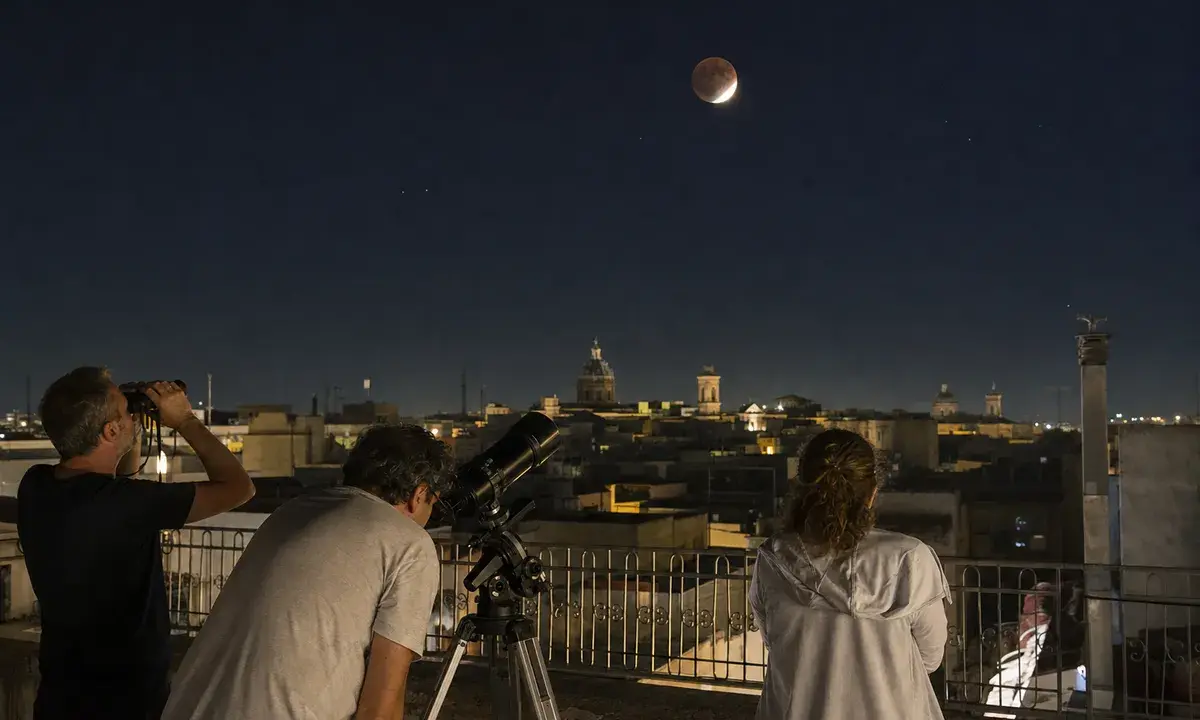Persone su una terrazza osservano la Luna parzialmente eclissata con binocolo e telescopio sopra i tetti della città