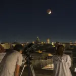 Persone su una terrazza osservano la Luna parzialmente eclissata con binocolo e telescopio sopra i tetti della città
