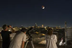 Persone su una terrazza osservano la Luna parzialmente eclissata con binocolo e telescopio sopra i tetti della città