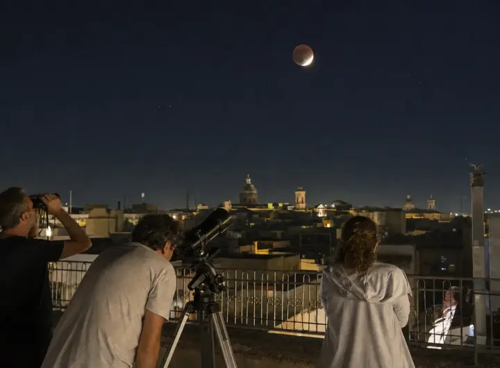 Persone su una terrazza osservano la Luna parzialmente eclissata con binocolo e telescopio sopra i tetti della città