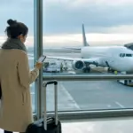 Viaggiatrice in aeroporto con trolley e smartphone davanti alle vetrate, con un aereo parcheggiato al gate