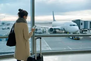 Viaggiatrice in aeroporto con trolley e smartphone davanti alle vetrate, con un aereo parcheggiato al gate