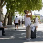 Donna beve acqua accanto a una valigia in un viale alberato; anziano seduto all’ombra durante una giornata estiva