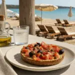 Frisella con pomodori, olive e capperi su tavolo vista spiaggia, con bottiglia d’olio e bicchiere d’acqua