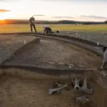 Scavo archeologico in un campo al tramonto con fossato curvo, paline di misura e ricercatori al lavoro