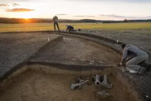 Scavo archeologico in un campo al tramonto con fossato curvo, paline di misura e ricercatori al lavoro