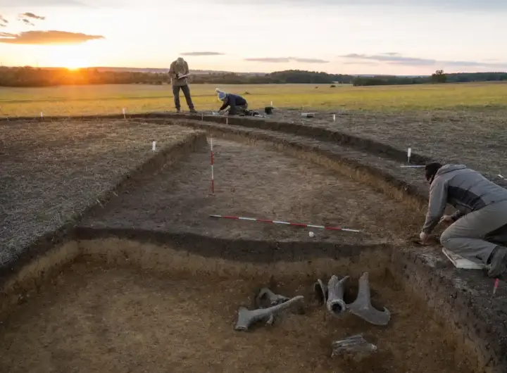 Scavo archeologico in un campo al tramonto con fossato curvo, paline di misura e ricercatori al lavoro