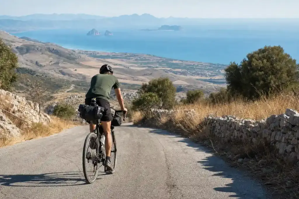 Ciclista con borse da viaggio pedala su una strada di montagna, con il mare e piccole isole sullo sfondo
