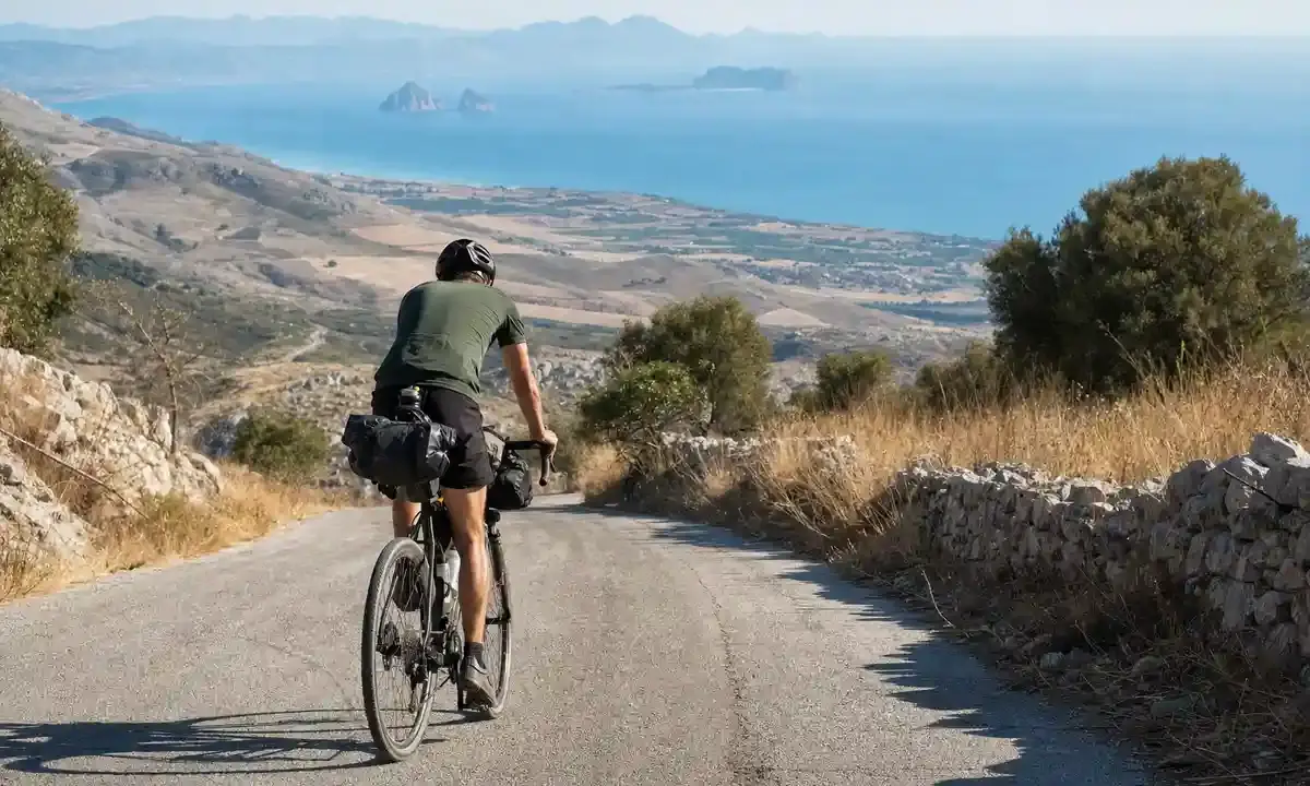 Ciclista con borse da viaggio pedala su una strada di montagna, con il mare e piccole isole sullo sfondo
