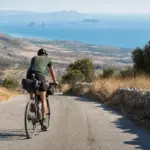 Ciclista con borse da viaggio pedala su una strada di montagna, con il mare e piccole isole sullo sfondo
