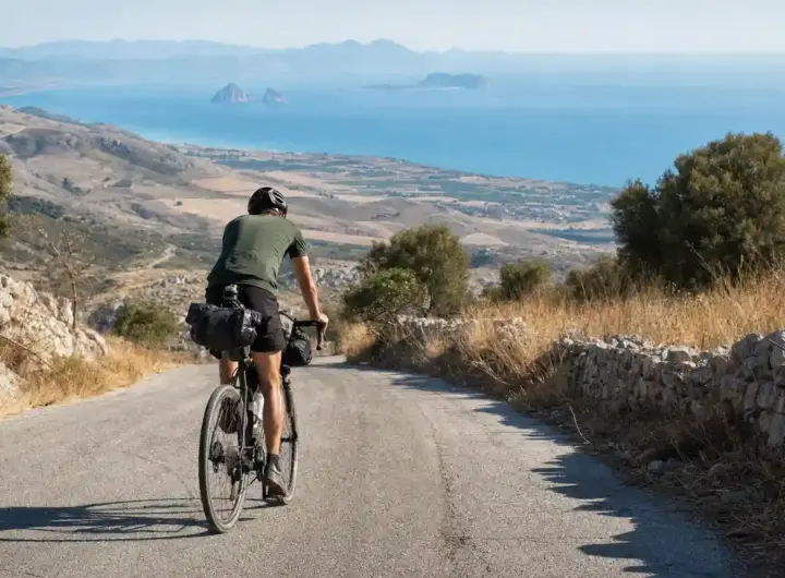 Ciclista con borse da viaggio pedala su una strada di montagna, con il mare e piccole isole sullo sfondo