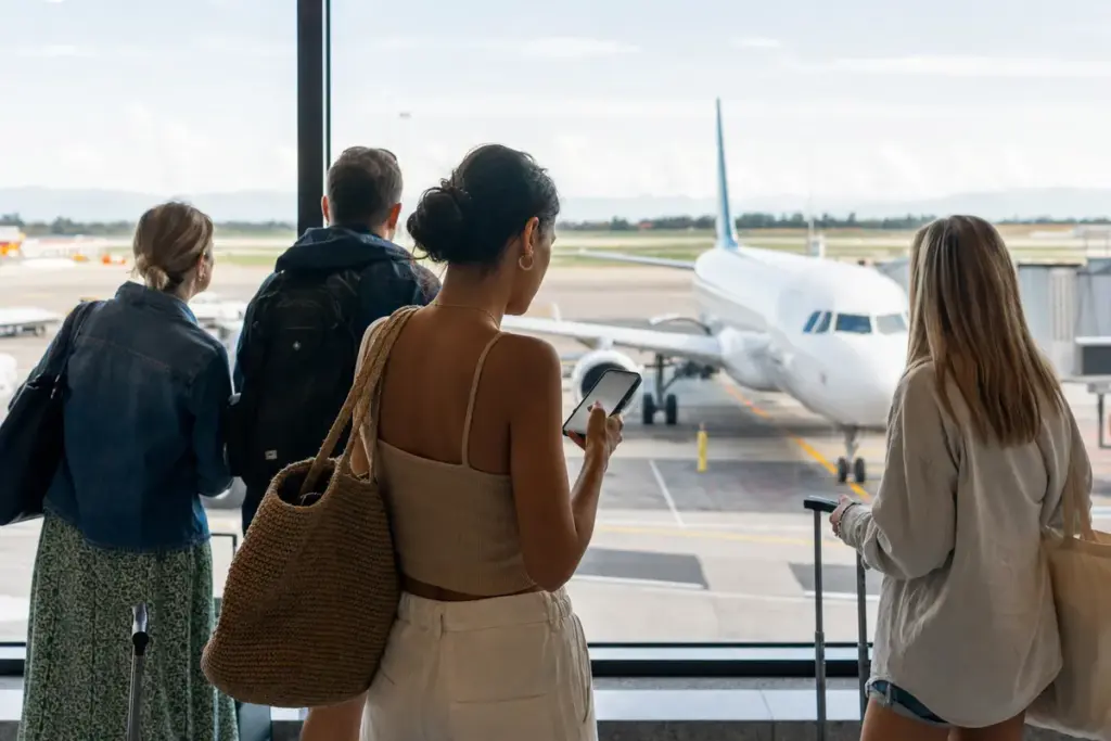 Viaggiatori con bagagli guardano un aereo in pista dal terminal, una donna controlla lo smartphone vicino alla vetrata