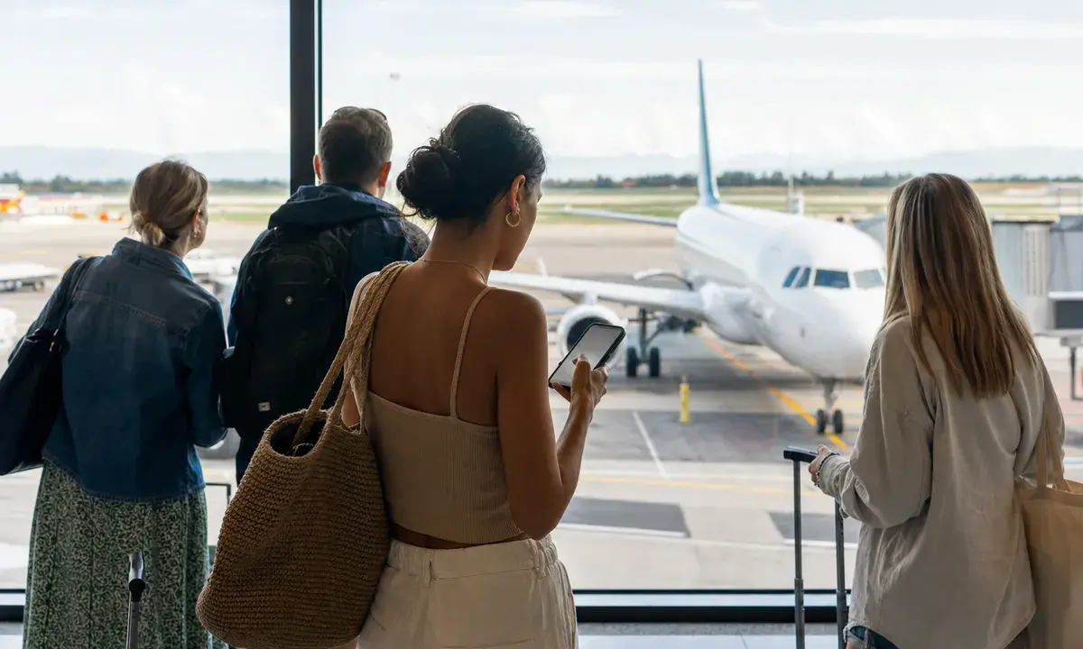 Viaggiatori con bagagli guardano un aereo in pista dal terminal, una donna controlla lo smartphone vicino alla vetrata