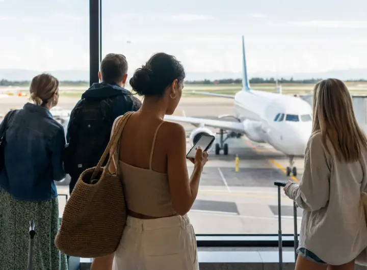 Viaggiatori con bagagli guardano un aereo in pista dal terminal, una donna controlla lo smartphone vicino alla vetrata