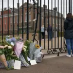 Mazzi di fiori e biglietti lasciati davanti ai cancelli di Kensington Palace, con visitatori sullo sfondo
