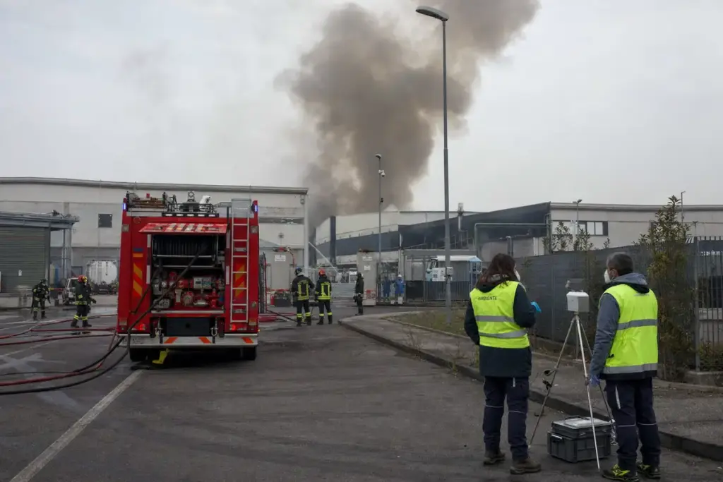 Autopompa e vigili del fuoco davanti a capannoni industriali con colonna di fumo, tecnici con strumentazione di monitoraggio