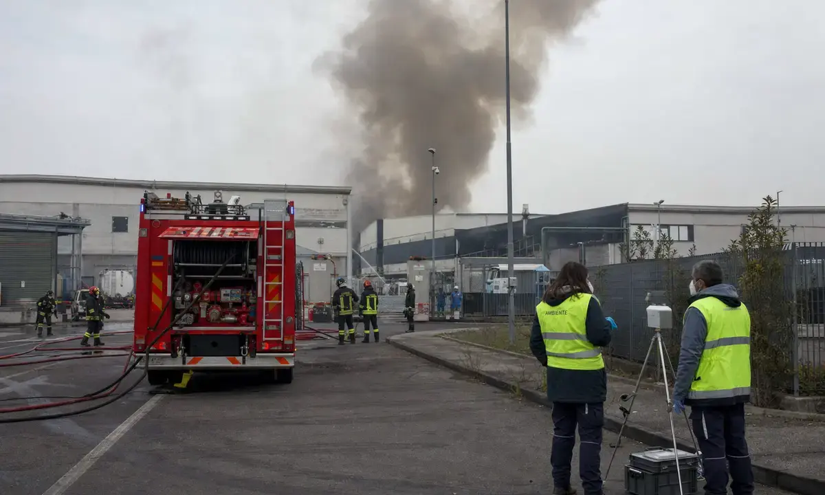 Autopompa e vigili del fuoco davanti a capannoni industriali con colonna di fumo, tecnici con strumentazione di monitoraggio