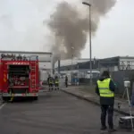 Autopompa e vigili del fuoco davanti a capannoni industriali con colonna di fumo, tecnici con strumentazione di monitoraggio
