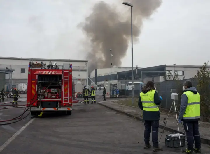 Autopompa e vigili del fuoco davanti a capannoni industriali con colonna di fumo, tecnici con strumentazione di monitoraggio