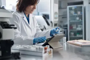 Tecnica di laboratorio in camice e guanti esamina una fiala e un tablet su un banco in un laboratorio ospedaliero