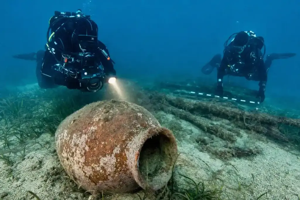 Subacquei documentano un grande dolium in terracotta sul fondale sabbioso, con posidonia e resti lignei sullo sfondo