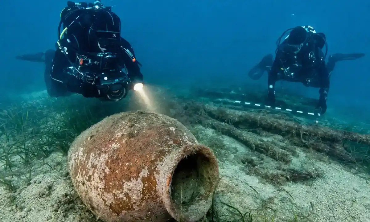 Subacquei documentano un grande dolium in terracotta sul fondale sabbioso, con posidonia e resti lignei sullo sfondo