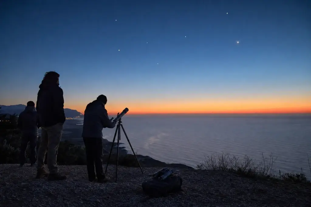 Osservatori con telescopio su un promontorio sul mare all’alba, con punti luminosi nel cielo azzurro