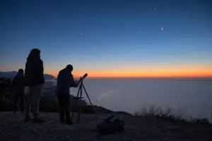 Osservatori con telescopio su un promontorio sul mare all’alba, con punti luminosi nel cielo azzurro