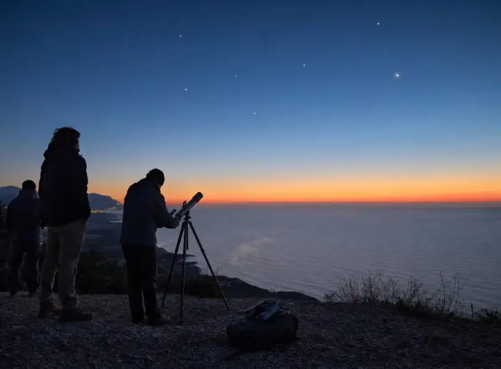 Osservatori con telescopio su un promontorio sul mare all’alba, con punti luminosi nel cielo azzurro