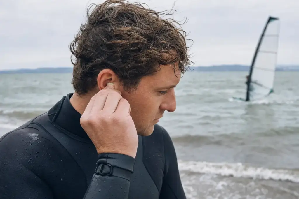Sportivo in muta che inserisce un tappo auricolare sulla riva, con acqua mossa e vela da windsurf sullo sfondo
