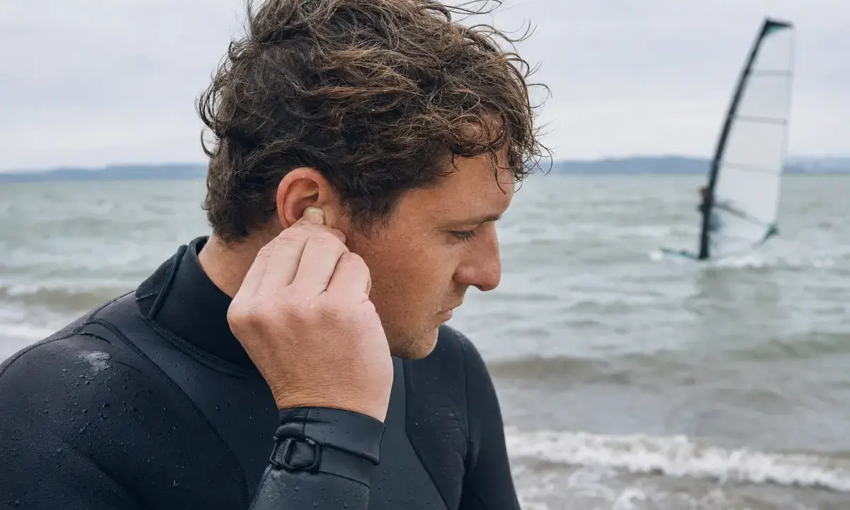 Sportivo in muta che inserisce un tappo auricolare sulla riva, con acqua mossa e vela da windsurf sullo sfondo