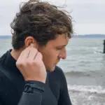 Sportivo in muta che inserisce un tappo auricolare sulla riva, con acqua mossa e vela da windsurf sullo sfondo