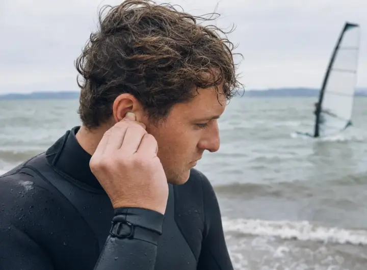Sportivo in muta che inserisce un tappo auricolare sulla riva, con acqua mossa e vela da windsurf sullo sfondo