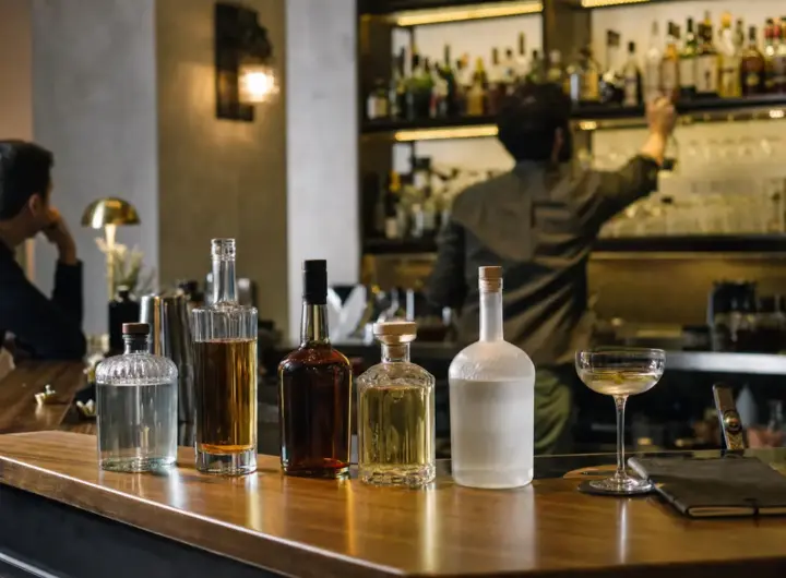 Bottiglie di spirits senza etichette su bancone in legno con cocktail e taccuino, bartender sullo sfondo