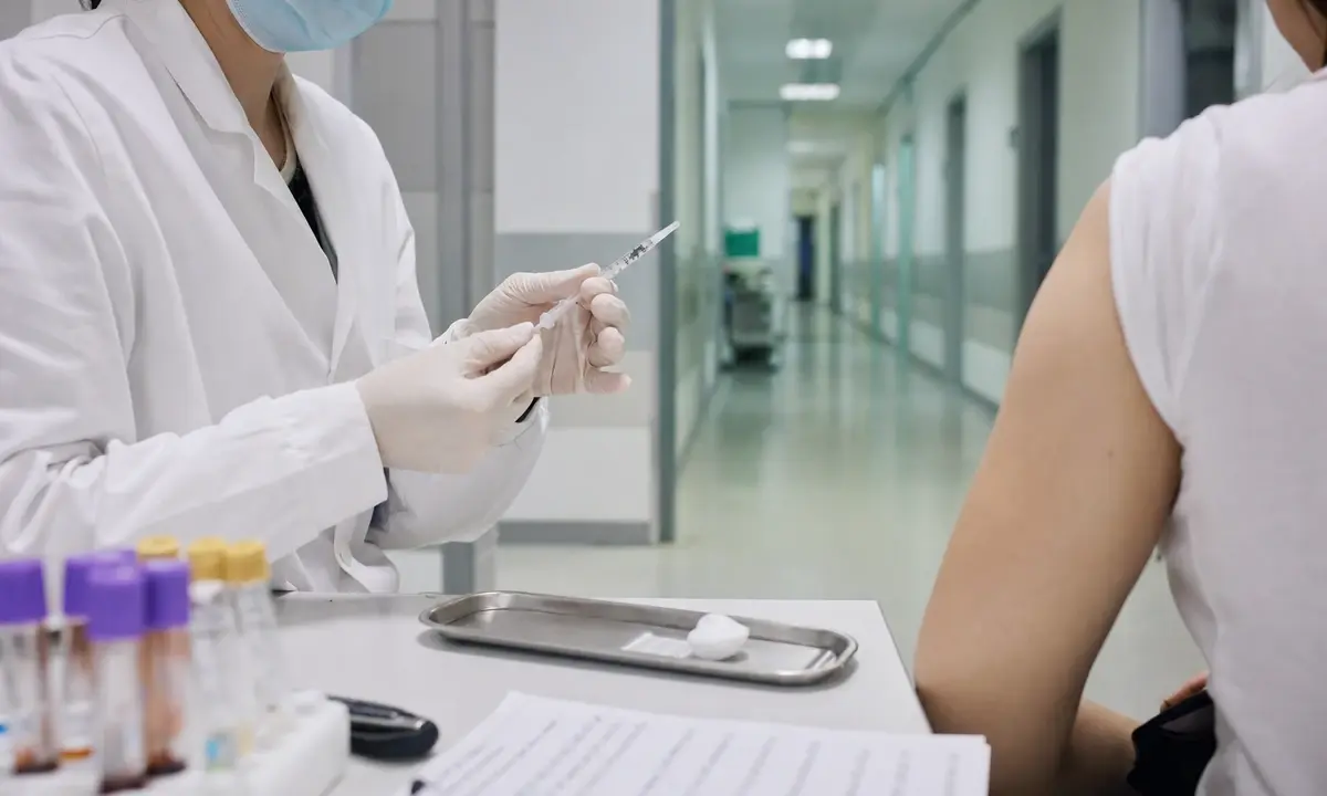 Operatore sanitario con mascherina prepara una siringa per un test, con paziente seduto e corridoio clinico sullo sfondo