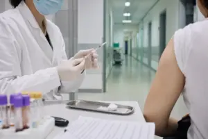 Operatore sanitario con mascherina prepara una siringa per un test, con paziente seduto e corridoio clinico sullo sfondo