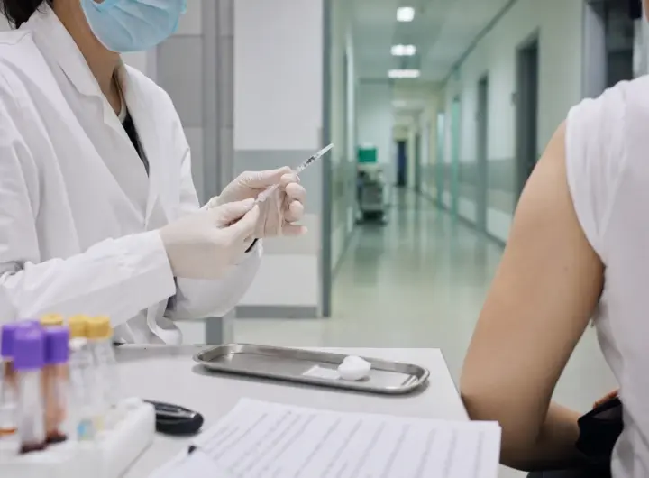 Operatore sanitario con mascherina prepara una siringa per un test, con paziente seduto e corridoio clinico sullo sfondo