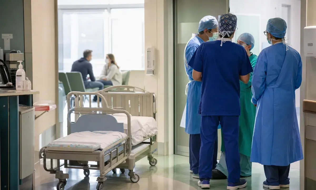 Corridoio ospedaliero pediatrico con lettino vuoto e personale sanitario in camice e mascherina che discute vicino a una