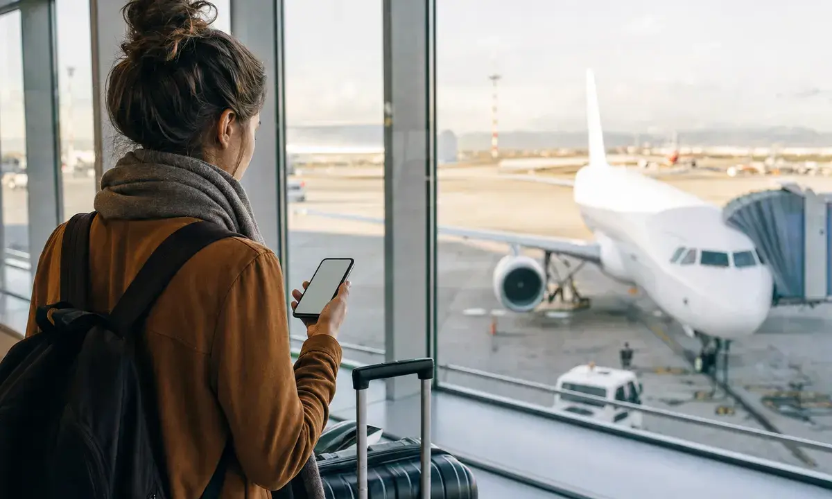 Viaggiatrice in aeroporto con smartphone e valigia davanti alle vetrate, con aereo parcheggiato sul piazzale