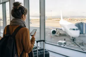 Viaggiatrice in aeroporto con smartphone e valigia davanti alle vetrate, con aereo parcheggiato sul piazzale