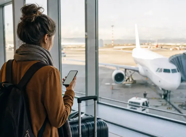 Viaggiatrice in aeroporto con smartphone e valigia davanti alle vetrate, con aereo parcheggiato sul piazzale