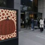 Poster con zucca a pois rossi davanti all’ingresso di un museo moderno, con visitatori in fila