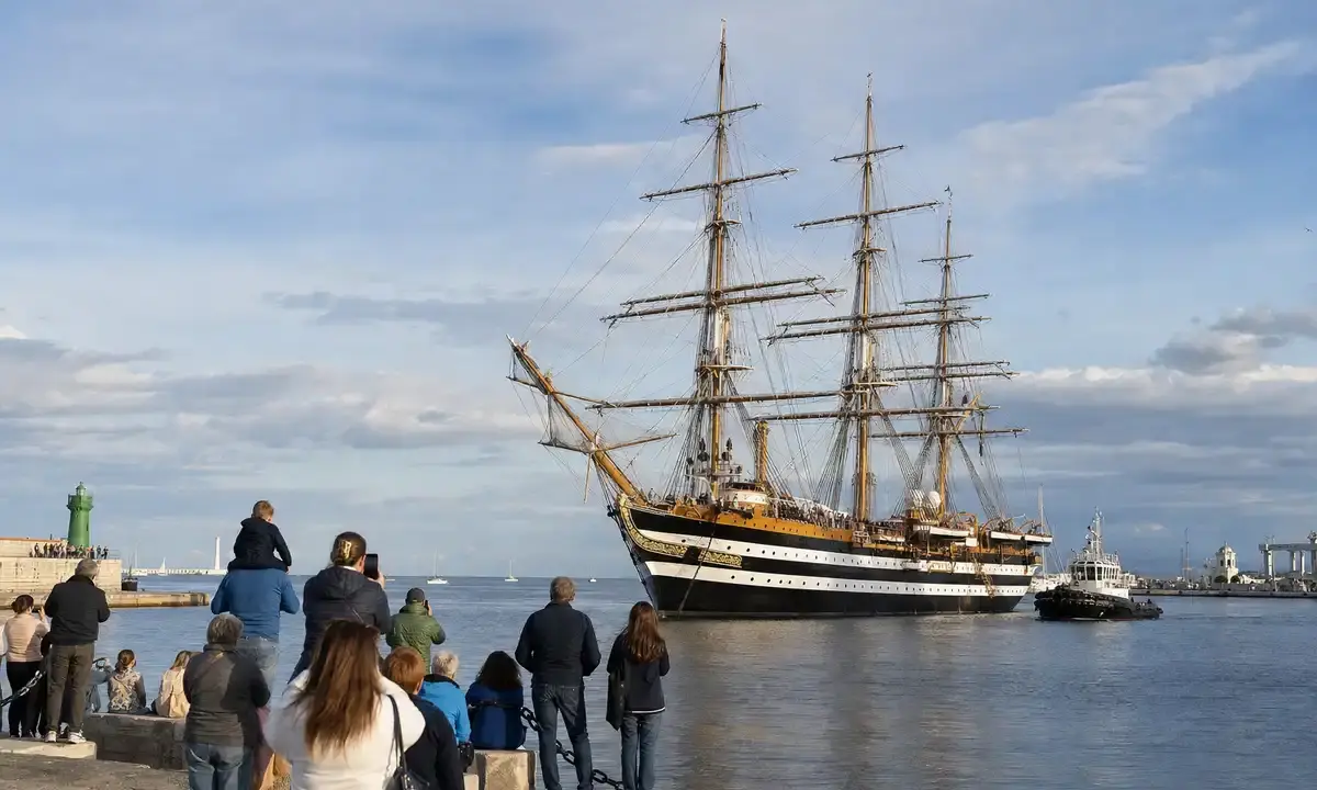 Nave a vela a tre alberi in porto, con persone sulla banchina che osservano l’arrivo